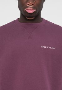 Purple sweatshirt made of soft cotton with ribbed neckline and cuff. Features white embroidered logo text "LYLE & SCOTT" on the front.