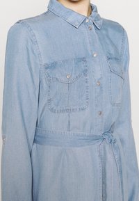 Ljusblå denimklänning med knäppning fram, två bröstfickor, långa ärmar och en knytbälte i midjan. Slät textur.