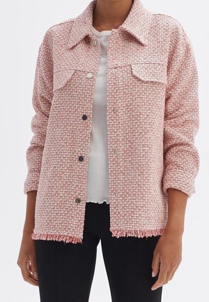 Frau trägt eine rosa-weiß strukturierte Knopfleistejacke mit Vordertaschen, ausgefranstem Saum, weißem Hemd darunter und schwarzen Hosen.