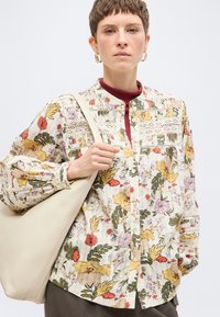 Γυναίκα με κοντά μαλλιά που φοράει μπλούζα με φλοράλ σχέδιο και μπορντό τοπ, κρατώντας μια μεγάλη μπεζ τσάντα ώμου σε απλό φόντο.