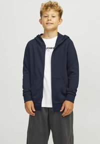 Felpa con zip color navy, con cerniera frontale, due tasche laterali e una consistenza morbida, indossata sopra una maglietta bianca, abbinata a pantaloni scuri.