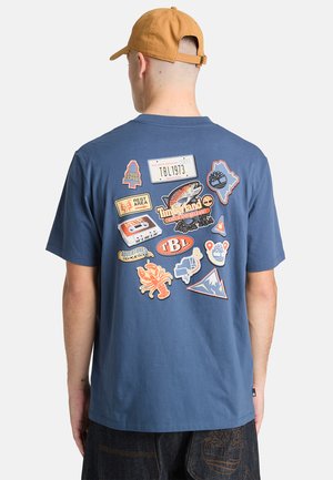 Maglietta di cotone blu, con diverse toppe colorate, tra cui grafiche di un pesce, alberi e montagne sul retro. Design casual.