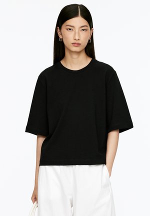 Schwarzes Baumwoll-T-Shirt mit Rundhalsausschnitt und kurzen Ärmeln. Das Design zeichnet sich durch einen entspannten Schnitt aus und wird mit weißen, weit geschnittenen Hosen kombiniert.