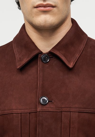 Άνδρας που φοράει σκούρο καφέ σουέντ μπουφάν με φαρδιά γιακά και δύο μεγάλα μαύρα κουμπιά κουμπωμένα μπροστά.