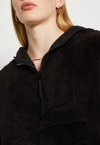 Donna che indossa una giacca in pile nera con zip e cappuccio, orecchini a cerchio d'oro e una sottile collana d'oro, mostrata dalla bocca al petto.