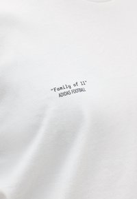 Λευκό ύφασμα με μαύρο τυπωμένο κείμενο που λέει "Οικογένεια των 11" και "ADIDAS FOOTBALL" με κεφαλαία γράμματα.