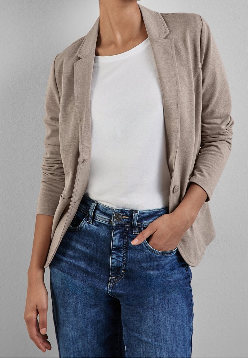 Femme portant un blazer beige par-dessus une chemise blanche et un jean bleu, une main dans la poche, sur un fond gris uni.