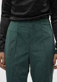 Pantalons côtelés teal à taille haute, avec des lignes verticales et une texture lisse, associés à un haut noir à manches longues.