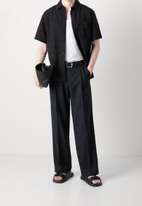 Korte zwarte textuurshirt over een witte tanktop, zwarte geplooide broek en zwarte sandalen; houdt een zwarte clutch in de hand.
