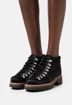 Bottes noires à la cheville, fabriquées en matière douce semblable à de la fourrure, avec des œillets et des lacets argentés, une semelle extérieure robuste texturée et une semelle intermédiaire en bois.