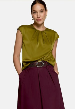 Femme portant un chemisier sans manches vert olive rentré dans un pantalon taille haute bordeaux avec une ceinture marron, mains dans les poches, et boucles d’oreilles créoles dorées.