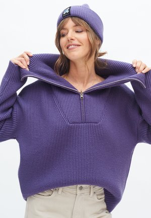 Lila gerippter Pullover mit halbem Reißverschluss am Kragen und einer Fronttasche. Getragen mit einer passenden lila Strickmütze, die mit einem Logo-Patch versehen ist.