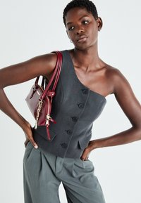 Gilet grigio a spalla unica con bottoni, abbinato a pantaloni larghi grigi. Gli accessori includono una piccola borsa in pelle bordeaux con dettagli in catena.