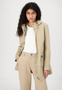 Giacca leggera beige impermeabile con cappuccio, zip frontale e tasche. Presenta una vita arricciata e una texture di tessuto liscia.