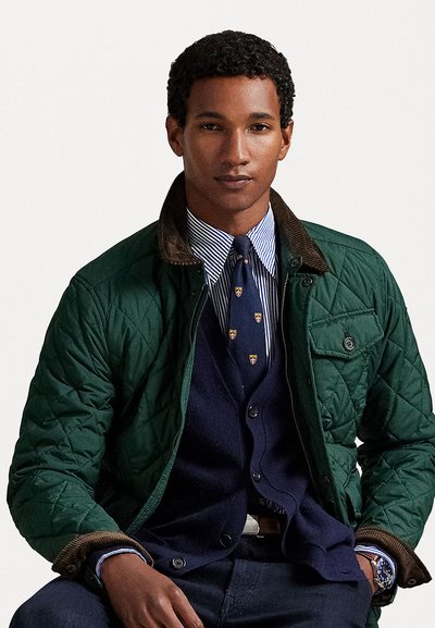 Chaqueta verde acolchada con un cuello de pana marrón, superpuesta sobre un cárdigan azul marino y una camiseta de rayas, con una corbata de estampado y jeans oscuros.