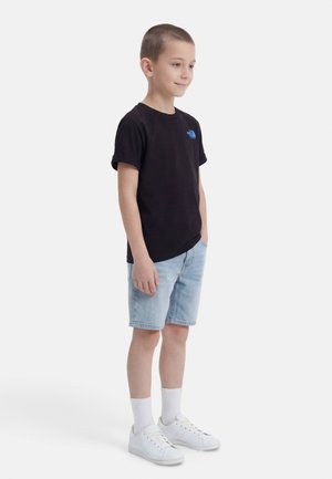 Ragazzo giovane in piedi con le mani nelle tasche, che indossa una maglietta nera, pantaloncini di jeans azzurri, calzini bianchi e scarpe da ginnastica bianche su sfondo bianco.