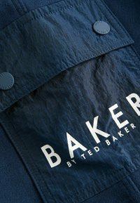 Temno modra tkaninska oznaka z mrežasto teksturo, z močnim belim besedilom "BAKER by TED BAKER" in dvema ujemajočima se okroglima gumboma na vrhu.