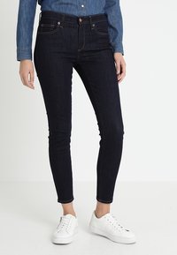 Vaqueros oscuros ajustados con cinco bolsillos, cierre frontal con botón y cremallera, y costuras en contraste. Combinados con zapatillas blancas y una camisa de mezclilla.
