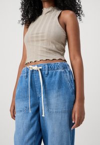 Beżowy prążkowany top crop z falbaniastym dołem i wysokim dekoltem, połączony z jasnoniebieskimi dżinsami z elastycznym pasem i białym sznurkiem.