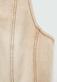 Material de denim bej cu cusături vizibile în fir mai deschis. Textura este netedă, cu un ușor luciu și o tăietură ascuțită la umeri.