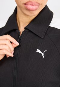 Svart jakke med glidelås, ribbet tekstur, og hvit Puma-logo. Kragen er fremtredende, med et kontrasterende hvitt panel på den ene siden.
