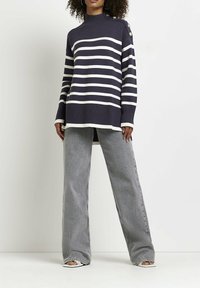 Femme portant un pull rayé bleu marine et blanc avec des boutons sur l'épaule, un jean gris à jambes larges, et des chaussures blanches à bout ouvert, debout contre un mur blanc.