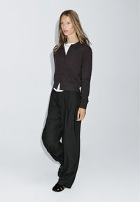 Cardigan marron foncé avec des boutons, porté sur une chemise blanche, associé à un pantalon large à rayures noires et à des chaussures noires brillantes.