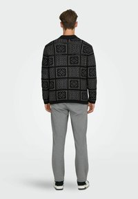 Svart mönstrad tröja med geometrisk design, bärs tillsammans med ljusgrå byxor. Tröjan har rund hals och ribbade muddar vid ärmarna.