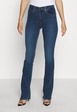 Woman wearing dark blue high-waist bootcut jeans and black open-toe heels standing against a plain background.
