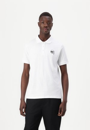 Polo bianco in cotone, con un'apertura a tre bottoni e un logo fumettistico nero sul lato sinistro del petto. Indossato con pantaloni neri.