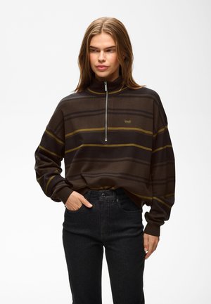 Jeune femme portant un pull marron rayé avec une fermeture éclair devant et un jean noir, debout avec une main dans la poche.