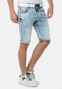 Helleblaue Jeansshorts mit ausgewaschenem Finish, hochgekrempelten Säumen und einem auffälligen Logo-Patch an der Vorderseite der Tasche. Getragen mit weißen Sneakern.