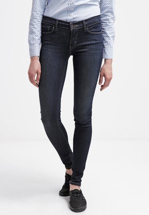 Jeans ajustados de denim azul oscuro con una textura suave. Presenta cinco bolsillos, un diseño de costura sutil y un ajuste ceñido. Combinados con zapatillas negras.