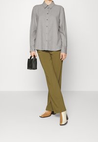 Camicia grigia a maniche lunghe con bottoni, pantaloni verde oliva e scarpe con tacco a blocco bicolore. Accessorio: piccola borsa nera. Design e texture semplici.