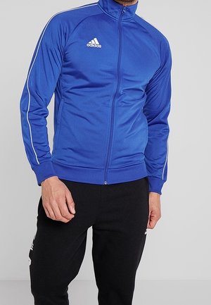 Mann, der eine blaue Adidas-Trainingsjacke mit Reißverschluss und weißen Streifen an den Ärmeln sowie schwarze Adidas-Hosen vor einem schlichten Hintergrund trägt.