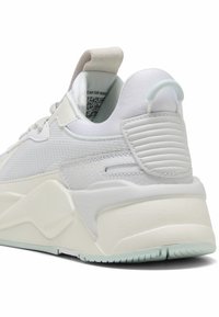 Bílé tenisky s texturovanou podrážkou, polstrovaným kotníkovým límcem a poutkem na patě, zobrazené ze zadního úhlu na bílém pozadí.