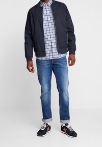 Giacca bomber blu navy sopra una camicia a quadri con tonalità di blu, verde e bianco. Jeans azzurri chiaro e sneakers nere con dettagli rossi completano il look.