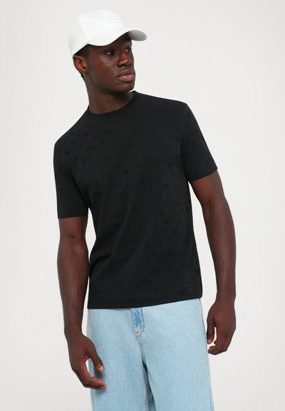 Junger Mann mit weißer Mütze, schwarzem, gemustertem kurzärmeligen Hemd und hellblauen Jeans, blickt zur Seite vor einem schlichten Hintergrund.