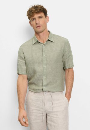 Mann trägt hellgrünes kurzärmeliges Hemd mit Knöpfen und beige Leinenhose mit Kordelzug, steht vor einem schlichten hellen Hintergrund.