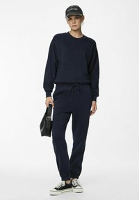Felpa blu navy e pantaloni jogger in tessuto morbido, con vita regolabile e polsini a coste, abbinati a sneakers nere e una borsa.