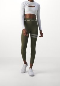 Top court à manches longues blanc avec des découpes, associé à des leggings verts olive avec des bandes réfléchissantes et une large ceinture. Chaussures de sport blanches.