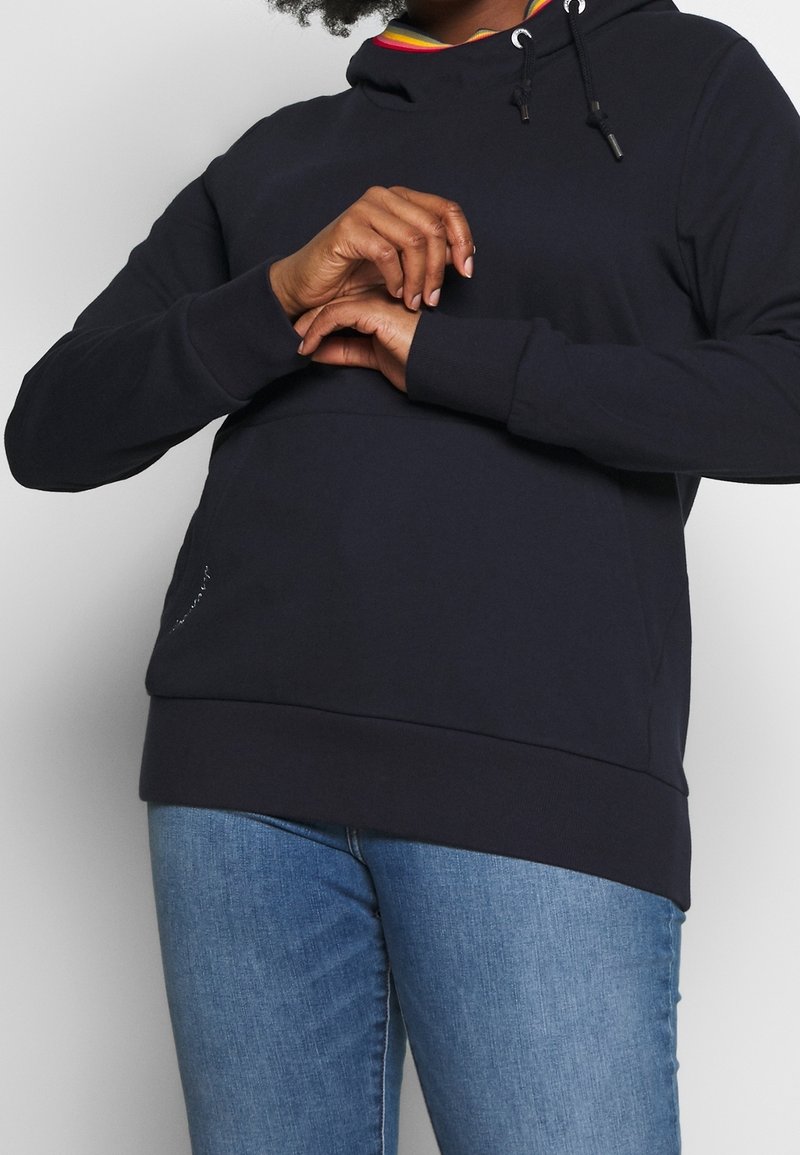 Personne portant un sweat à capuche noir avec une doublure de capuche à rayures arc-en-ciel et un jean bleu clair, mains jointes au niveau de la poitrine sur un fond uni.