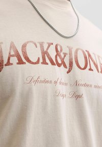 Μπεζ βαμβακερό μπλουζάκι με κόκκινο τυπωμένο κείμενο "JACK & JONES" και "Definition of denim Nineteen ninety". Διαθέτει στρογγυλή λαιμόκοψη και ομαλή υφή.