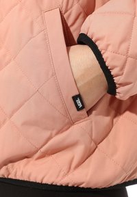 Handen instoppad i sidofickan på en vadderad rosa Vans-jacka med svarta elastiska manschetter och svart trim i nederkant.