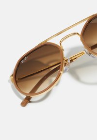 Ochelari de soare aviator maro cu accente metalice aurii, având lentile cu gradient și un cadru gros din plastic maro. Include garnituri nazale transparente.