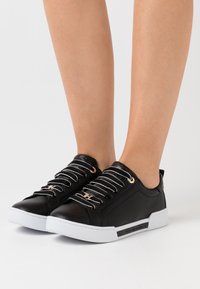 Person, der schwarze Leder-Low-Top-Sneaker mit weißen Sohlen und weißen gestreiften Schnürsenkeln trägt und vor einem schlichten hellen Hintergrund steht.