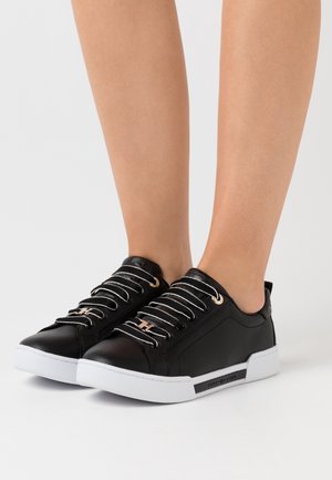 Persona che indossa sneakers nere in pelle a basso profilo con suole bianche e lacci a strisce bianche, in piedi su uno sfondo chiaro e uniforme.
