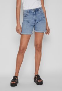 Calças de ganga cortas em azul claro, com bainhas dobradas, design clássico de cinco bolsos e fecho com zíper. Combinadas com sandálias de plataforma pretas.