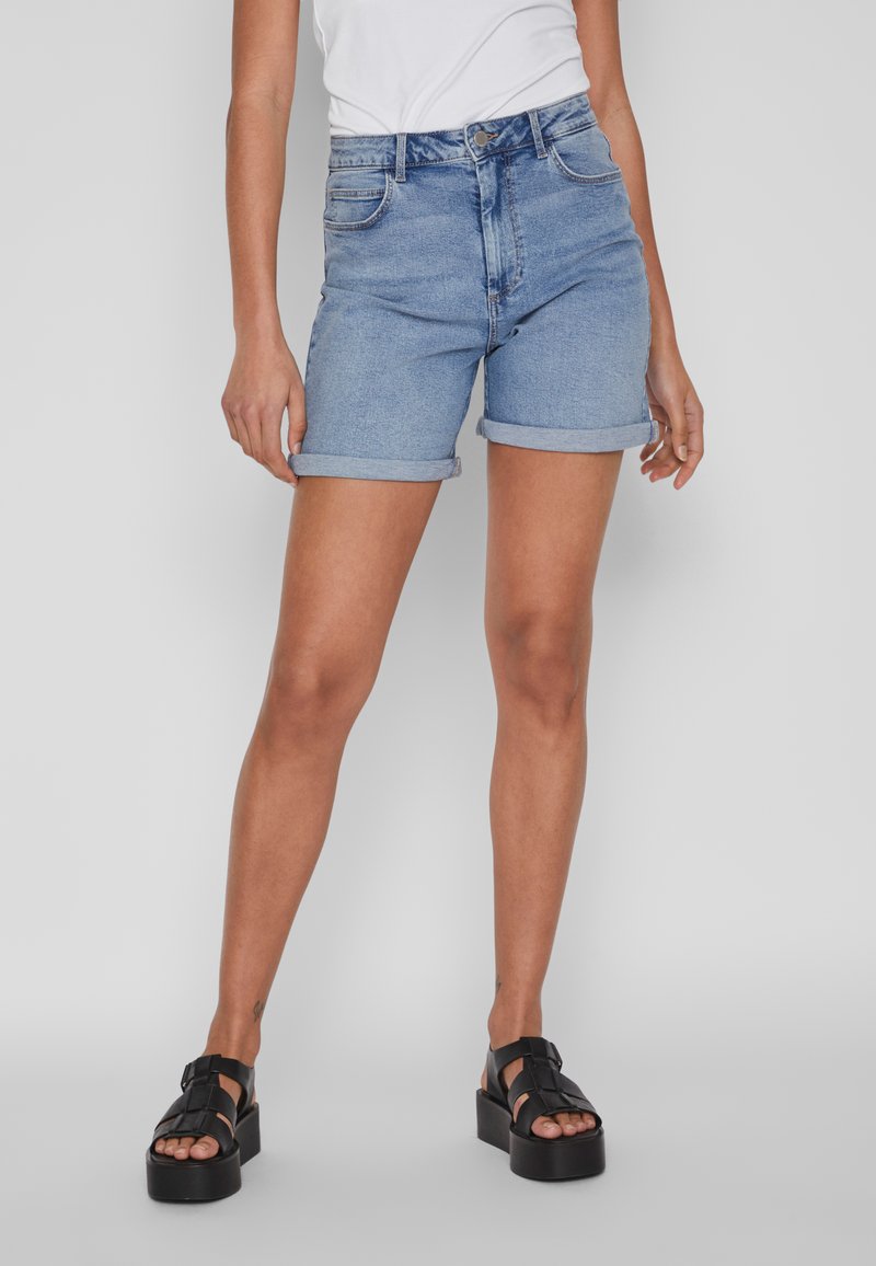 Calças de ganga cortas em azul claro, com bainhas dobradas, design clássico de cinco bolsos e fecho com zíper. Combinadas com sandálias de plataforma pretas.