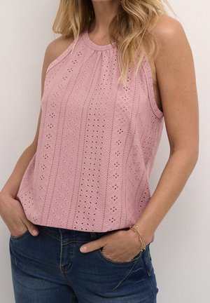 Kobieta w różowym topie bez rękawów z wzorem ażurowym i niebieskich dżinsach, z rękami w kieszeniach, nosząca złotą bransoletkę łańcuchową.